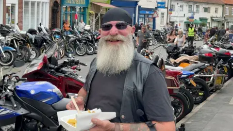 A person with a long white beard stands among rows of parked motorcycles at a busy biker gathering, holding a tray of chips in a bustling town street.