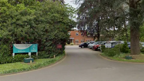 Google The entrance to a red-bricked building. There is a blue and white sign for the hospice on the left.