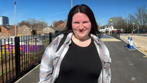 A young woman with dark hair wearing a black top and checked shirt smiles at the camera. She is standing on a pavement next to black metal railings at what appears to be a newly built railway station. Behind her, colourful hoarding, street lights and terraced houses are visible under a clear blue sky.
