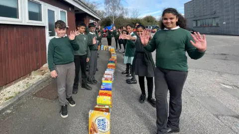 Outdoors, children standing beside a line of cereal boxes on the floor, which are standing up ready to be pushed.