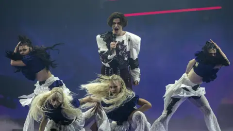 Marko Bosnjak holding a microphone and singing with his mouth wide during his Eurovision performance. He is surrounded by four female dancers with very long swishing hair - two are blonde and two are dark. The dancers are wearing white lacy flared trousers and ruffled navy blue crop tops. Bosnjak has mid-length dark hair and is wearing a long-sleeved black and white lacy top with huge shoulder pads.