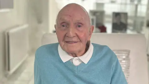 An elderly man wearing a light blue jumper over a white dress shirt. He is smiling at the camera.