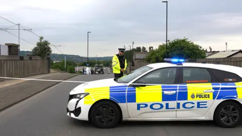 Oli Constable A police officer stands next to a police car and tape at the scene on Woodburn Road. Forensic officers can be seen in the background.