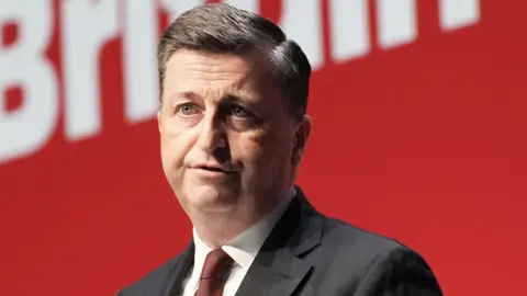 PA Media Douglas Alexander, who has dark hair, wearing a white shirt, red tie and dark suit. He is sitting in front of a red background. 