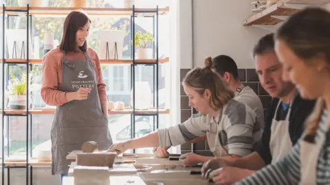 Handout Annabel Cusack, wearing a grey apron, stands with a group of people who are creating their own pottery. The group are sitting with clay modelling items and one woman is reaching for some clay. The pottery studio has white walls and grey tiles and a stand in the window showing products that have been made.