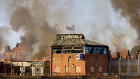 Media News GNP Ltd A derelict, smoke-damaged building. The roof has fallen in with the fire and all the windows are blown out. The bottom floor windows are bricked up and there appears to be a "for auction" sign on the side of the building. Flames can be seen rising from the top floor, with smoke pouring out, as an aerial fire officer directs a hose into the flames.
