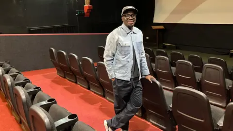 A man with a black hat, wearing a blue denim jacket and black cargo pants is standing inside a cinema screen. The carpet is red and the seats are black, with a white screen in the background.
