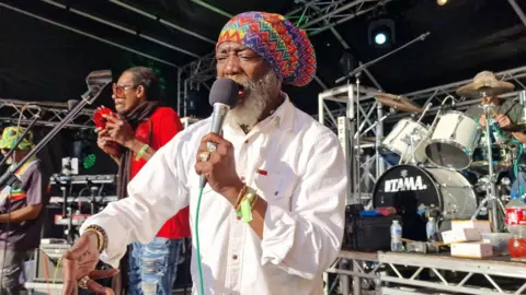 The Vanlife Festival A man is wearing a multicoloured rasta cap and a white shirt. He is signing into a microphone on a stage. In the background, a man is playing the tamborine and another is playing the drums.