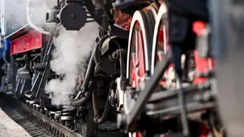 Getty Images A generic and close up view of a train's wheels on train tracks. Steam can be seen leaving a part of the train. Some of the machinery is painted red.