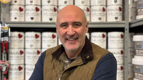 A man with a bald head and grey-coloured stubble smiles at the camera. He is wearing a green gilet and a blue top. He is standing in front of tins of paint.