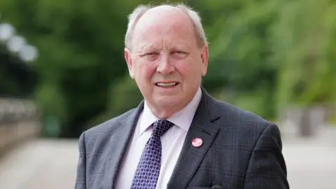 PA Media Jim Allister is pictured outside Stormont. He is wearing a dark grey check suit, white shirt, blue patterned tie and a small red badge. 