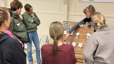Victoria Gemma Photography Sarah Bolger talking about fuses during the taster sessions. She is pointing at different ones that are put on a wooded board. Six women are around her, listening. Three of them have their backs to the camera.