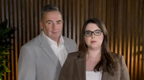 John Hunt, in a grey blazer, and Amy, who wears dark-rimmed glasses and has long brown hair and wears a brown blazer
