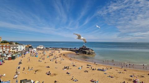 Seagulls fly above beach-goers at Broadstairs in Kent, with a calm sea, and a blue sky with wispy clouds