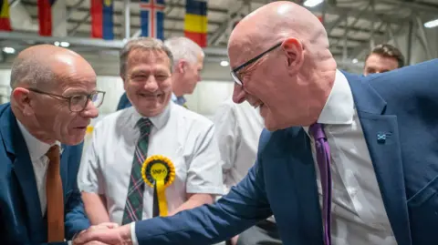 John Swinney shakes a man's hand. They are standing in front of a smiling man wearing an SNP rosette