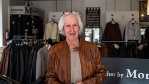 James Morgan wearing a brown leather jacket stands inside a clothing shop, with racks of jackets and garments displayed in the background. A pair of glasses rests on the person's head, and a shop sign is partially visible behind the counter.