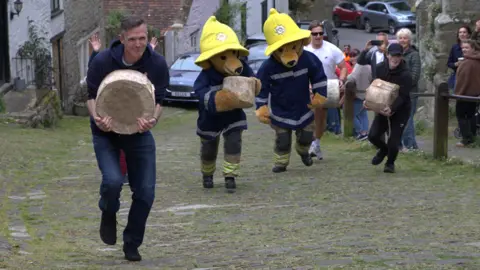 Two men flanking two people dressed in teddy bear costumes, who are dressed as firefighters, running up the hill. 
