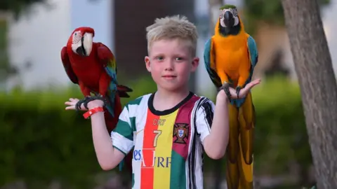 Family Louie on holiday. He is wearing a sports top and has a parrot perched on each hand.