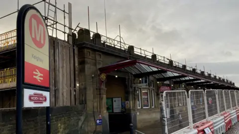 A red and yellow sign with black lettering spelling out Keighley and another sign underneath with Worth Valley written in white letters on a maroon background. There is white metal fencing and the canopied entrance to the station in the background.   