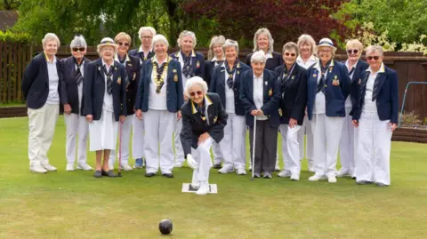Contributed A group of older woman stand on a bowling green together and smile at the camera. One woman in front of them crouches slightly as she bowls a black ball in front of her. They wear matching white trousers, blue blazers and navy ties. Some wear white bowler hats.