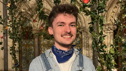 Choir member Max Ryan pictured at the chapel. He is a young man and is wearing a denim shirt, denim dungarees and a blue and white neck-tie. He is smiling widely at the camera