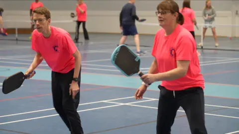 Jamie Niblock/BBC Sonia and her doubles partner, standing side by side, wearing their pink Suffolk Punch Pickleball T-shirts. They are both action-ready, with knees slightly bent, mid match, holding their pickleball paddles.