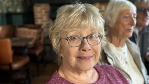 Jenny Barnard at an Age UK event at the Hop Blossom pub in Farnham, Surrey. Jenny is wearing glasses and a pink jumper.