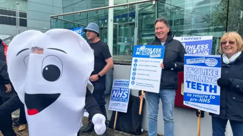 Connor Bennett/BBC More campaigners outside Endeavour House during a protest. One person is dressed in a costume as a tooth with a face on it.