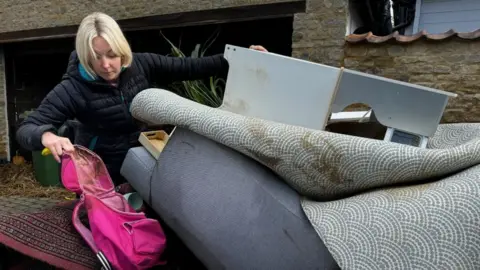 Emma Bilton with medium-length blond hair wearing a blue sweater tips up a waterlogged sofa in her house and picks up a pink child's bag from the floor