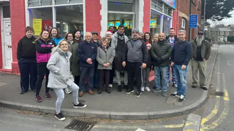A large group of around twenty people of mixed ages stand smiling on a street corner outside the red-brick ‘Victoria Fish Bar.’ The blue shop sign is above them, and the group is gathered on the pavement in front of the takeaway, with houses and a traffic-calmed street visible in the background. Several people are wearing winter coats and a few festive hats.