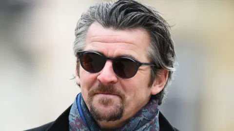 Joey Barton with goatee beard, black sunglasses, a Paisley scarf and a black coat.