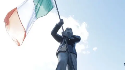 Close shot of a bronze Bobby Sands statue holding and orange white and green Irish flag against a blue sky.