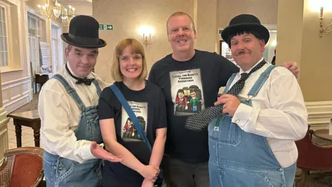 Dean Sills Rebecca and Dean Sills pose for a photo with two Laurel and Hardy lookalikes. The father and daughter smile and wear black t-shirts, while the performers wear Demin dungarees, white shirts and ties.