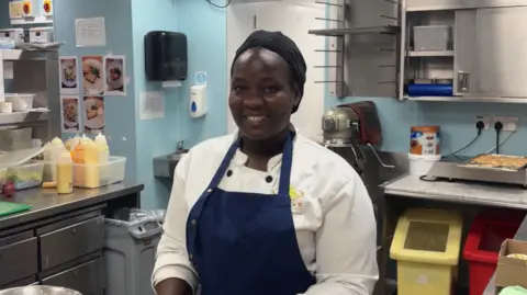 Lady wearing chefs jacket and blue apron in a kitchen smiling at the camera.