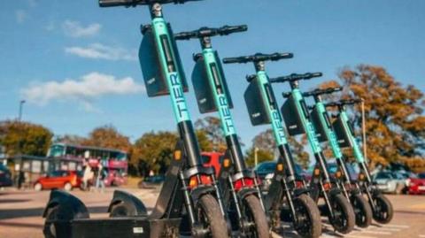 A row of teal coloured e-scooters in a parking bay with blurred bus stop and road in the background