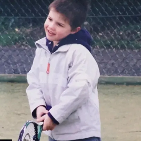 Stanford University A young boy wearing a coat stands on a tennis court holding a racket.