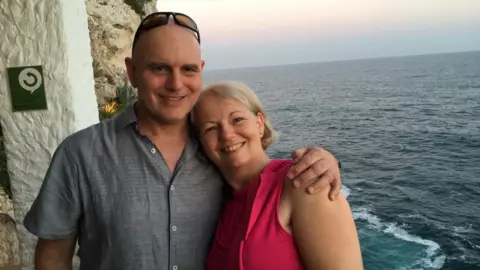 Mark Butler with his arm around his wife. They are stood next to an old brick wall with the sea behind them. 