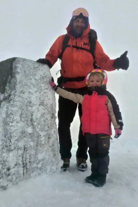 Craig Moffat Faye Moffat and her dad Craig smiling as they touch the concrete trig point on the summit of Ben Nevis which is covered in snow and ice. It is snowy and foggy. They are both wearing waterproof winter gear. Faye is in a bright pink jacket with beige and black sleeves. The hood is up over the top of the hood of a multi-coloured fleece. She is also wearing grey trousers, black boots and winter gloves. Craig is wearing an orange jacket with black trousers, gloves, and boots. He has ski goggles on the top of his head and a rucksack on his back. He is giving a thumbs-up sign.