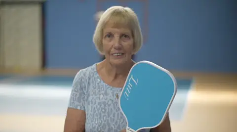 Gill Jeffrey standing on a pickleball court at Hingham Sports Centre in Norfolk. She is looking directly at the camera and smiling. She has a short blonde bob with a fringe and is wearing a pale blue leopard print T-shirt and is holding a blue pickleball racket.