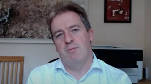 Graeme Downie is wearing a blue shirt and sitting in an office in front of a chair, printer and pictures on a wall.