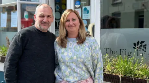 Ryan Marsland and a colleague stood directly outside their shop. They are both looking at the camera and are smiling. There are window boxes with greenery in front of the large glass windows which has the company's name and logo on.
