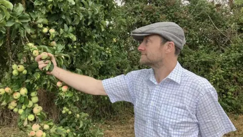 The image shows Barney Butterfield wearing a grey flat cap and short sleeved checked shirt, holding an apple still attached to the branch of a tree. 