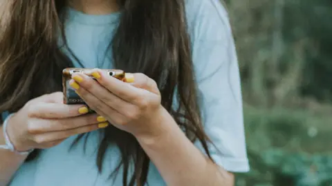 Girl with long dark hair, wearing a blue top looks down at her phone