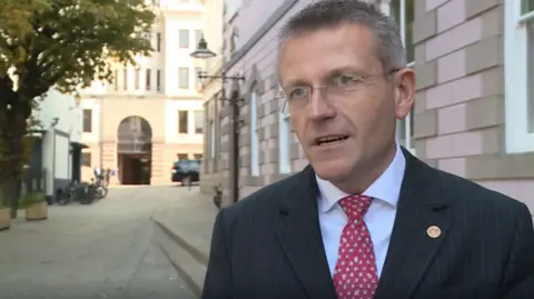 Philip Ozouf stood on the right wearing a suit. He has a pinstriped blazer with a red and white tie. He is looking to the left and is talking. In the background are buildings, one with an arched doorway.