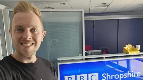 Ian Handley Ian has strawberry blonde hair styled in a quiff and he is taking a selfie in front of a blue and white BBC Radio Shropshire sign