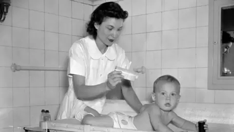 Getty Images A black and white photograph of a woman, who is dressed in a white dress and holding talcum powder, while a baby in a nappy is laying on their stomach.