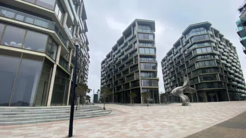 Three tower blocks in the Horizon development in St Helier. A black street light is on a pathway surrounding the tower blocks and a statue that looks like a sea wave is also on the path.