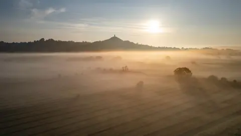 A view of the valley with Glastonbury Tor in the distance. It is sunrise and fog is rising from the valley, ulluminated by the sun.