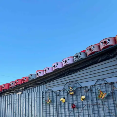 Tom Hicks A dozen Henry hoover faces, of different colours, sit on top of a blue shed. Attached to the shed is a wire trellis with sculptures of bees and butterflies attached. 