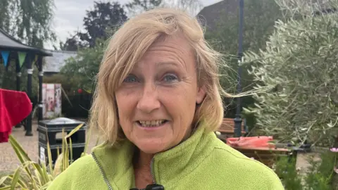 John Devine/BBC A headshot of Sandie Mellors who has sandy-coloured, shoulder length hair parted to her right. She is wearing a lime green fleece top. There are shrubs, bushes and a black litter bin in the background. 